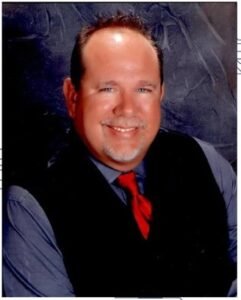 Smiling man named Wes DeVault with short brown hair and a goatee, wearing a dark vest, blue shirt, and red tie, posed in front of a textured dark background.